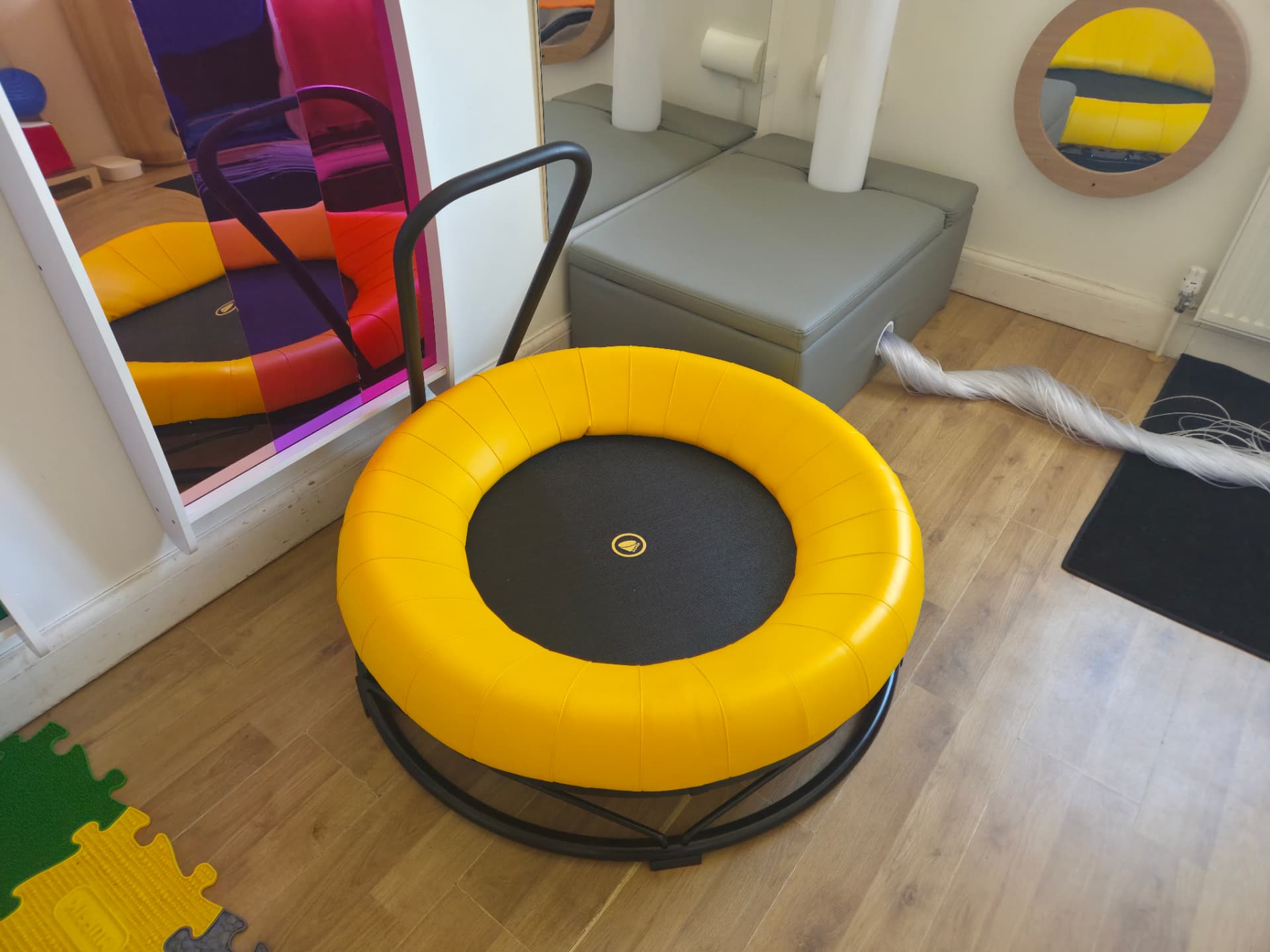 A small padded trampoline with a support handle in the sensory room, used for movement and proprioceptive input